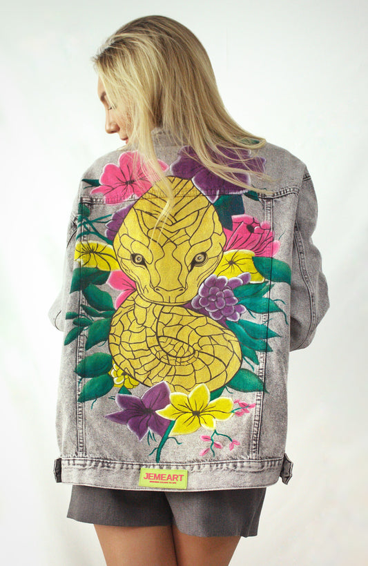  Hand-painted denim jacket with a golden cobra design on the back, surrounded by vibrant flowers in fuchsia, violet, yellow, and green leaves. Distressed texture and matching floral details on the front, designed for women who embrace art and individuality.